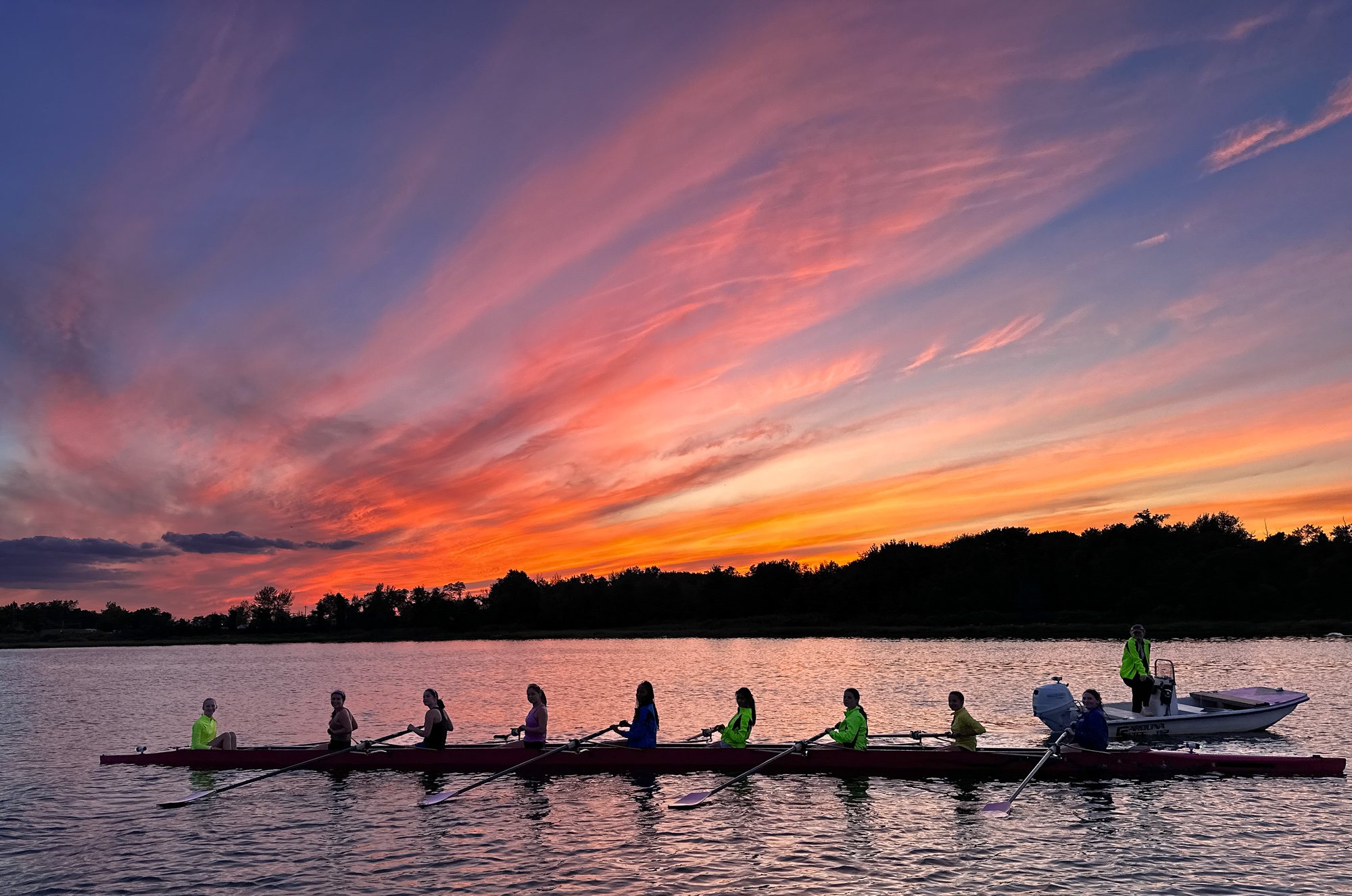 A CIR eight silhouetted against a blazing pink and orange sunset, oars resting on glass-calm water